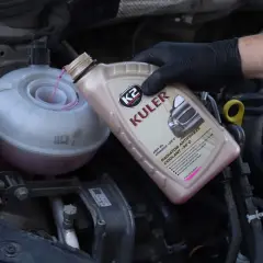 Main d'une personne portant un gant noir verse du liquide de refroidissement rose dans le réservoir d'un moteur de voiture.