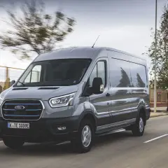 Un Ford Transit gris roule sur une route, sa hauteur imposante se démarquant dans le paysage urbain.