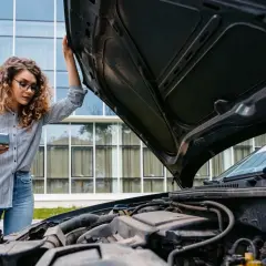 Femme cherchant sur son téléphone où trouver le code moteur sur carte grise, capot ouvert.