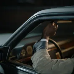 Main tenant de son volant qui grince, un homme en costume regarde par la fenêtre de sa voiture, pensif.