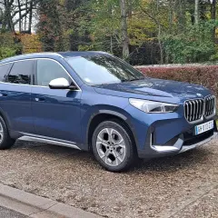 Avis sur la BMW X1 sDrive 18d 150 ch : ce SUV bleu est garé sur un chemin pavé, entouré d'arbres aux couleurs d'automne.