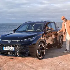 Un homme en tenue décontractée pose à côté d'un Volkswagen T-Roc noir, le moteur prêt à sillonner la côte.