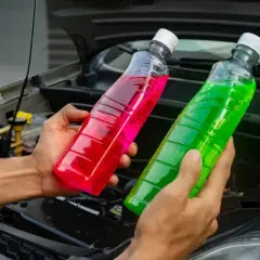 Deux bouteilles de liquide de refroidissement, une verte et une rose, sont tenues devant le moteur d'une voiture. Quel liquide de refroidissement vert pour quelle voiture ?