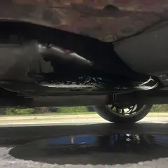 Sous la voiture, une flaque sombre indique une fuite d'essence. Le réservoir et le pot d'échappement sont visibles.