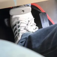 Un pied en basket Adidas appuie sur la pédale de frein molle. Le tapis de sol rouge est visible.