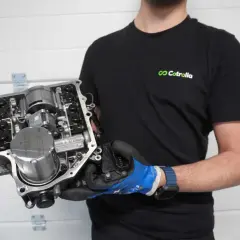 Un homme en t-shirt noir tient un ensemble de transmission complexe, montrant le levier de vitesse et d'autres composants mécaniques.