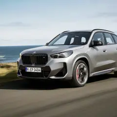 Un essai BMW X1 gris argenté roule sur une route côtière, l'océan bleu en arrière-plan.