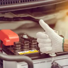 Main d'une personne portant un gant blanc fait un pouce levé près d'une batterie de voiture. Une astuce pour le démarrage à froid diesel.
