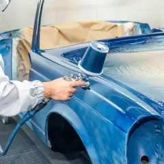 Un peintre automobile, protégé, applique une couche de peinture bleue sur une voiture classique. Une garantie réparation auto assure la qualité du travail.