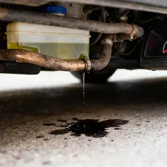 Gouttes de liquide de refroidissement tombent d'un tuyau sous une voiture, formant une flaque sombre sur l'asphalte. Le tableau de bord affiche un voyant d'alerte.