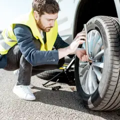 Un homme en gilet jaune s'apprête à changer un pneu crevé sur le bord de la route. Le cric est déjà en place.