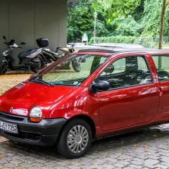 Twingo 1 rouge garée sur pavés, avec des scooters et des arbres en arrière-plan. Pensez à vérifier le filtre habitacle Twingo 1 pour un air pur.