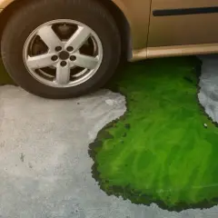 Une flaque de liquide de refroidissement vert vif s'étend sur le sol en béton sous une voiture dorée, indiquant un niveau liquide de refroidissement trop bas.