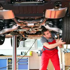 Un mécanicien examine le dessous d'une voiture sur un pont élévateur, lampe torche à la main. Il vérifie les composants pour s'assurer que tout est en ordre, comme lors d'un contrôle technique.