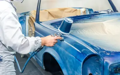 Un peintre automobile, protégé, applique une couche de peinture bleue sur une voiture classique. Une garantie réparation auto assure la qualité du travail.