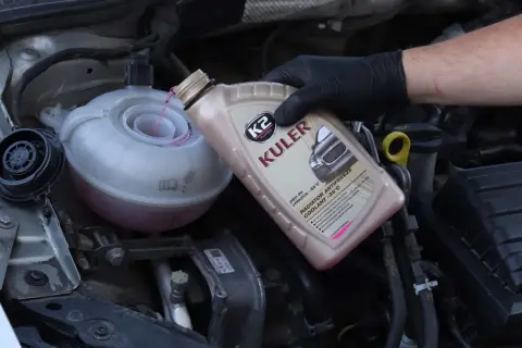 Main d'une personne portant un gant noir verse du liquide de refroidissement rose dans le réservoir d'un moteur de voiture.
