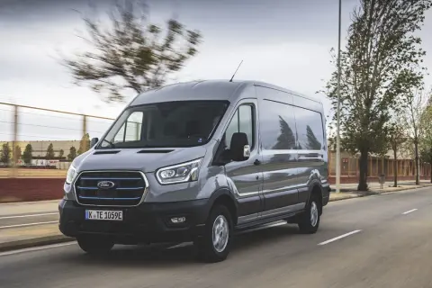 Un Ford Transit gris roule sur une route, sa hauteur imposante se démarquant dans le paysage urbain.