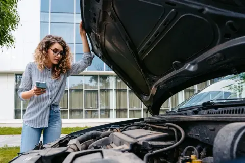 Femme cherchant sur son téléphone où trouver le code moteur sur carte grise, capot ouvert.