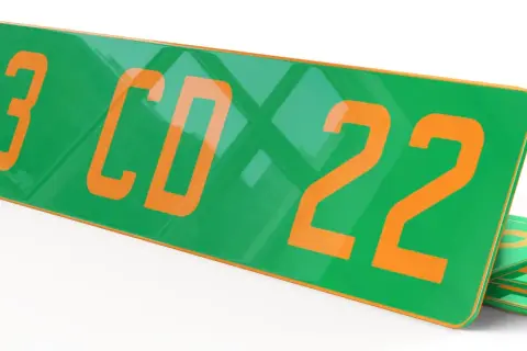 Plaque verte et orange avec les numéros "123 CD 22" en gros caractères. D'autres plaques similaires sont empilées en dessous.