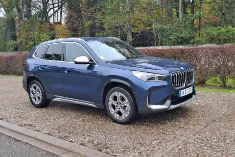 Avis sur la BMW X1 sDrive 18d 150 ch : ce SUV bleu est garé sur un chemin pavé, entouré d'arbres aux couleurs d'automne.