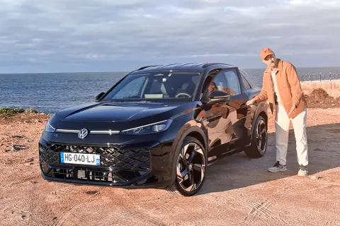 Un homme en tenue décontractée pose à côté d'un Volkswagen T-Roc noir, le moteur prêt à sillonner la côte.