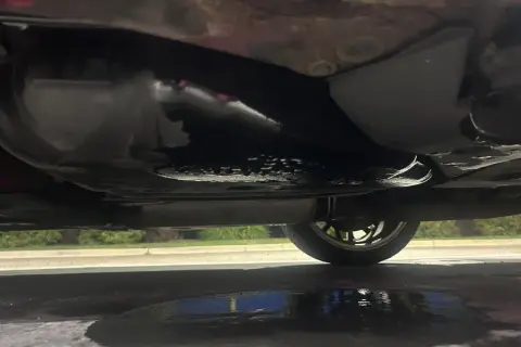Sous la voiture, une flaque sombre indique une fuite d'essence. Le réservoir et le pot d'échappement sont visibles.