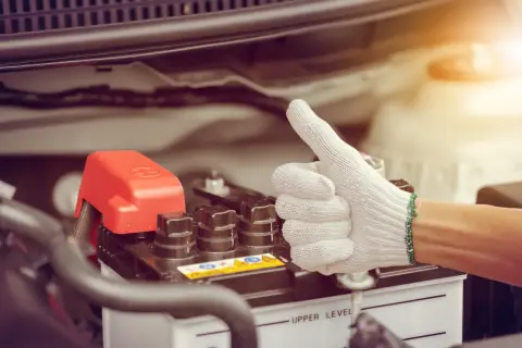 Main d'une personne portant un gant blanc fait un pouce levé près d'une batterie de voiture. Une astuce pour le démarrage à froid diesel.