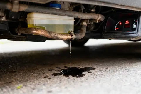Gouttes de liquide de refroidissement tombent d'un tuyau sous une voiture, formant une flaque sombre sur l'asphalte. Le tableau de bord affiche un voyant d'alerte.