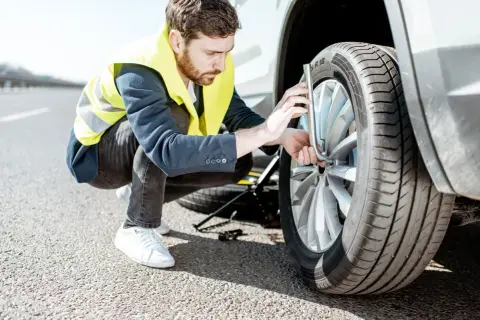 Un homme en gilet jaune s'apprête à changer un pneu crevé sur le bord de la route. Le cric est déjà en place.