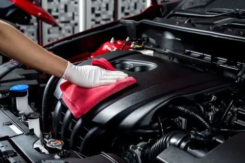 Main d'une personne portant un gant blanc, essuyant le capot du moteur d'une voiture avec un chiffon rouge pour nettoyer l'huile moteur.