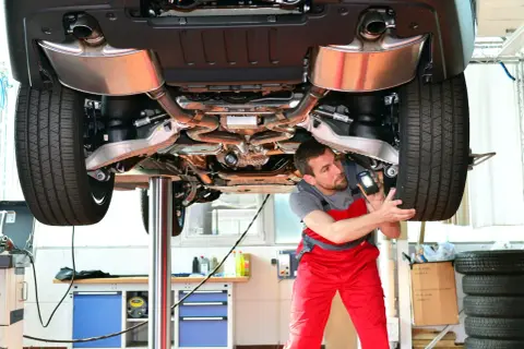 Un mécanicien examine le dessous d'une voiture sur un pont élévateur, lampe torche à la main. Il vérifie les composants pour s'assurer que tout est en ordre, comme lors d'un contrôle technique.