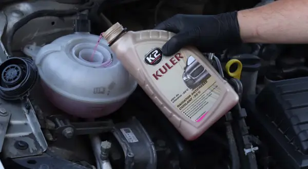 Main d'une personne portant un gant noir verse du liquide de refroidissement rose dans le réservoir d'un moteur de voiture.
