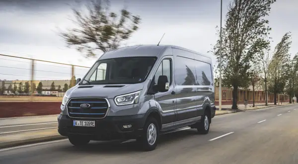 Un Ford Transit gris roule sur une route, sa hauteur imposante se démarquant dans le paysage urbain.
