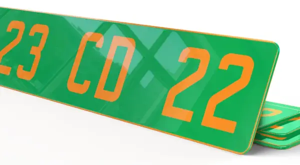 Plaque verte et orange avec les numéros "123 CD 22" en gros caractères. D'autres plaques similaires sont empilées en dessous.