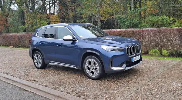 Avis sur la BMW X1 sDrive 18d 150 ch : ce SUV bleu est garé sur un chemin pavé, entouré d'arbres aux couleurs d'automne.