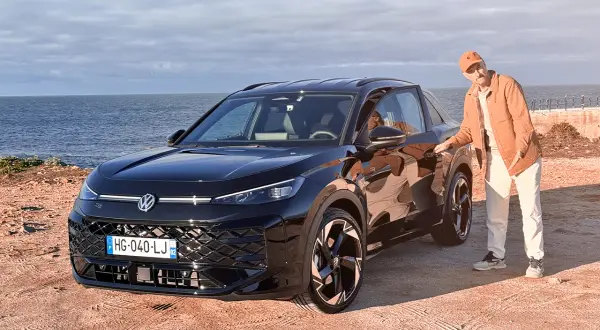 Un homme en tenue décontractée pose à côté d'un Volkswagen T-Roc noir, le moteur prêt à sillonner la côte.