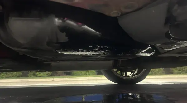 Sous la voiture, une flaque sombre indique une fuite d'essence. Le réservoir et le pot d'échappement sont visibles.
