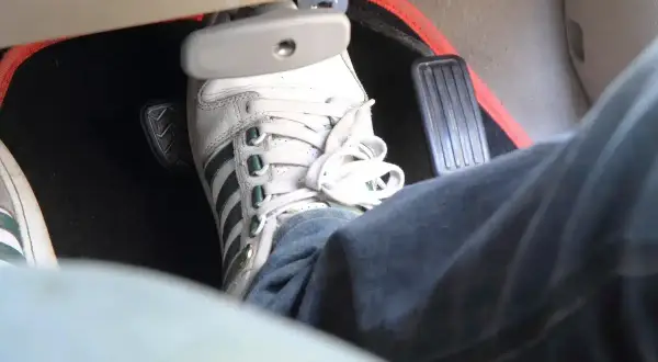 Un pied en basket Adidas appuie sur la pédale de frein molle. Le tapis de sol rouge est visible.
