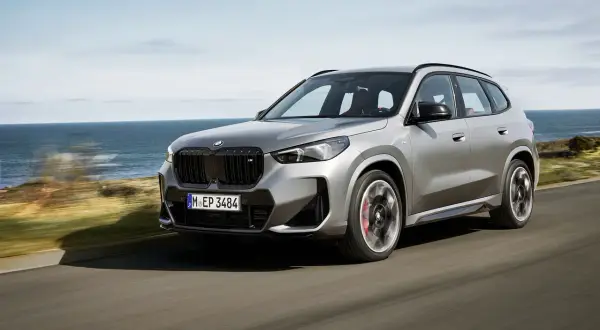 Un essai BMW X1 gris argenté roule sur une route côtière, l'océan bleu en arrière-plan.