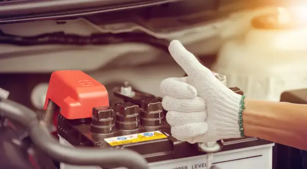 Main d'une personne portant un gant blanc fait un pouce levé près d'une batterie de voiture. Une astuce pour le démarrage à froid diesel.