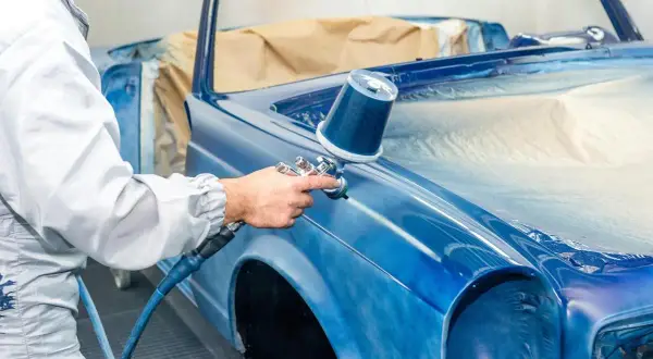 Un peintre automobile, protégé, applique une couche de peinture bleue sur une voiture classique. Une garantie réparation auto assure la qualité du travail.