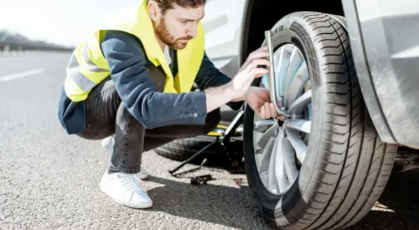 Un homme en gilet jaune s'apprête à changer un pneu crevé sur le bord de la route. Le cric est déjà en place.