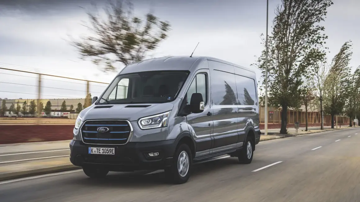Un Ford Transit gris roule sur une route, sa hauteur imposante se démarquant dans le paysage urbain.
