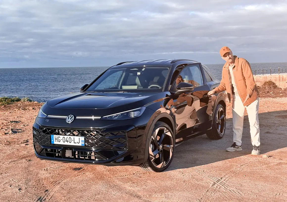 Un homme en tenue décontractée pose à côté d'un Volkswagen T-Roc noir, le moteur prêt à sillonner la côte.