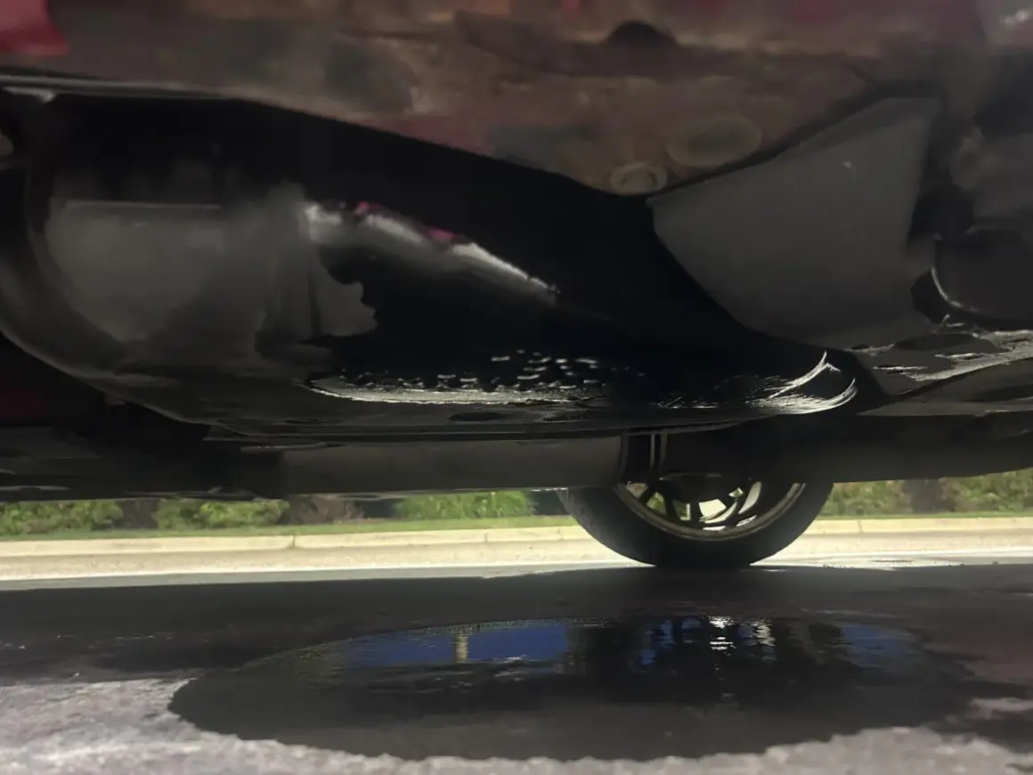 Sous la voiture, une flaque sombre indique une fuite d'essence. Le réservoir et le pot d'échappement sont visibles.
