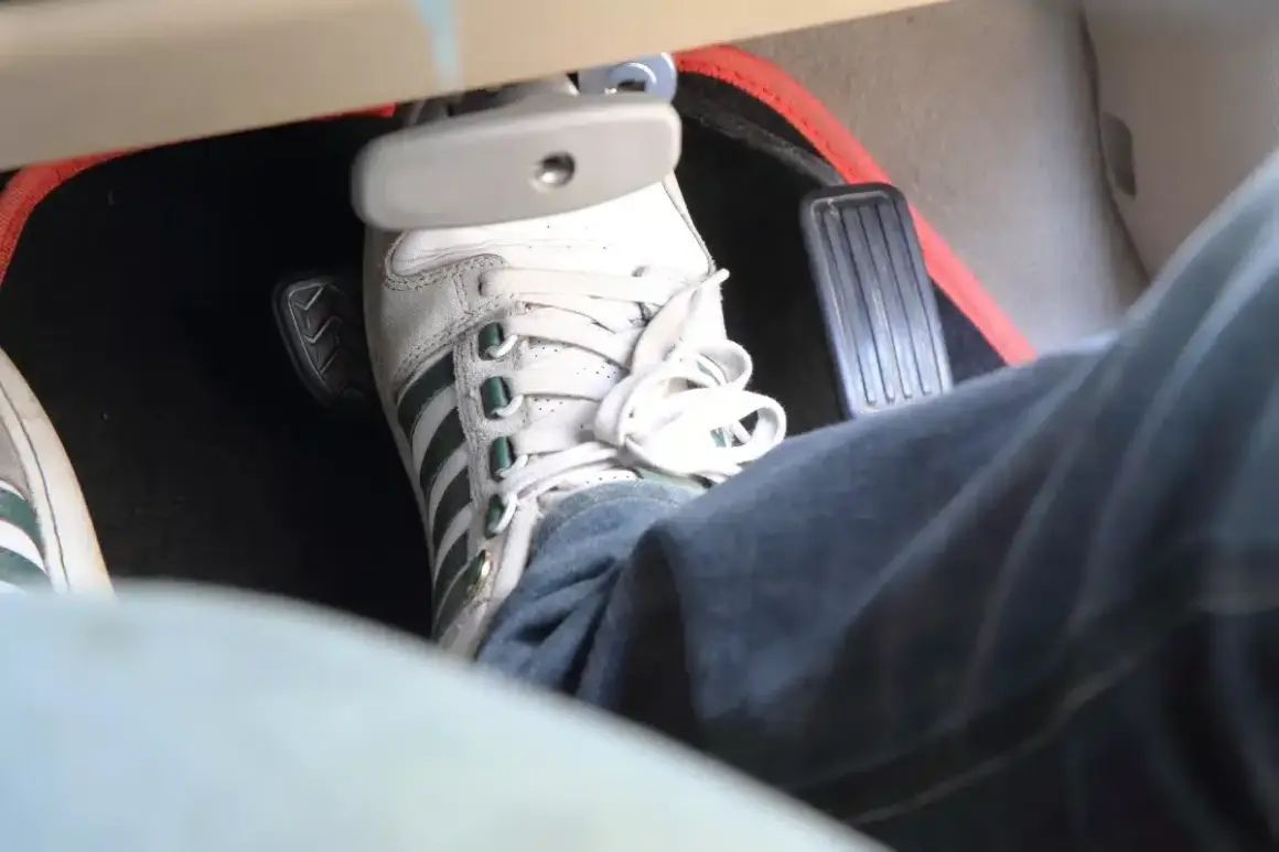 Un pied en basket Adidas appuie sur la pédale de frein molle. Le tapis de sol rouge est visible.