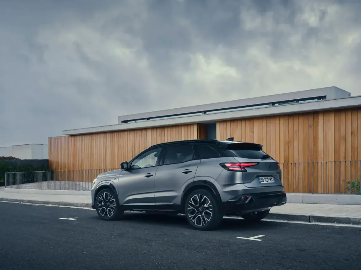 Vue arrière d'un SUV gris Renault Austral, stationné sur une route devant un bâtiment moderne en bois. La motorisation est performante.