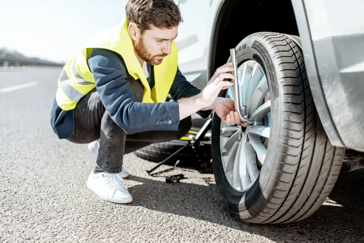 Un homme en gilet jaune s'apprête à changer un pneu crevé sur le bord de la route. Le cric est déjà en place.