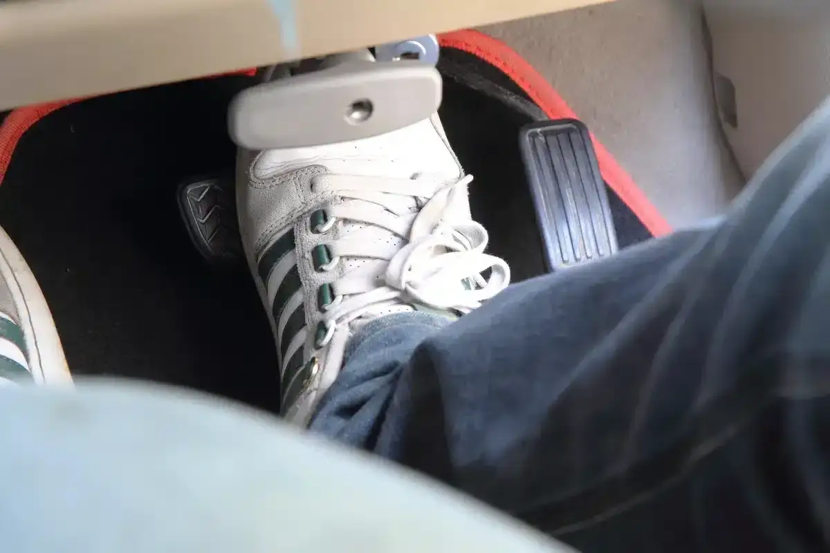 Un pied en basket Adidas appuie sur la pédale de frein molle. Le tapis de sol rouge est visible.