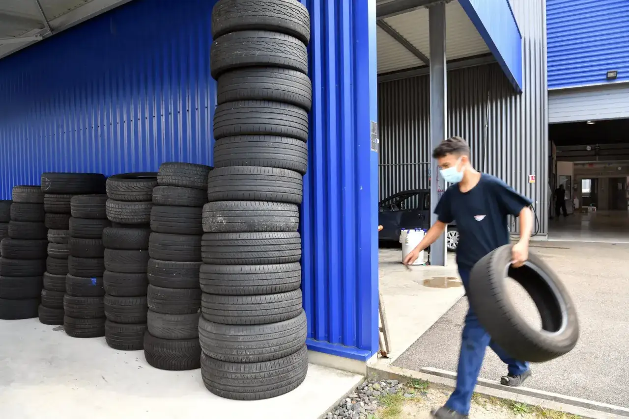 Un jeune homme masqué transporte un pneu usagé, prêt à le jeter. Des piles de pneus s'alignent le long d'un mur bleu.